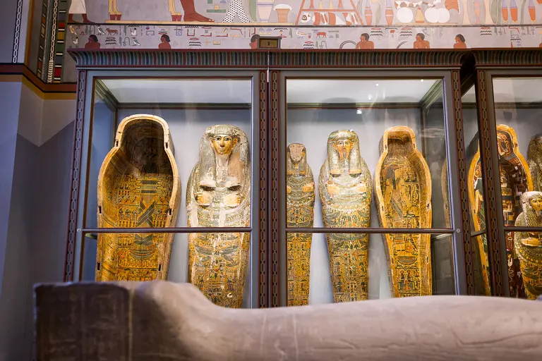 In einem Ausstellungsraum sind mehrere antike mumifizierte Särge in Vitrinen zu sehen. Die Särge sind bunt bemalt und zeigen verschiedene Details, die die ägyptische Kunst und Kultur repräsentieren. Im Vordergrund befindet sich ein Teil einer Couch.