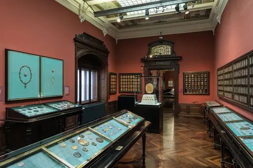 Das Bild zeigt einen museumsausstellungsraum mit Vitrinen, die Schmuckstücke und Münzen präsentieren. Die Wände sind in einem warmen Rotton gehalten, und die Vitrinen sind auf eleganten Holztischen angeordnet, während ein großer Rahmen mit einer runden Uhr in der Mitte steht.