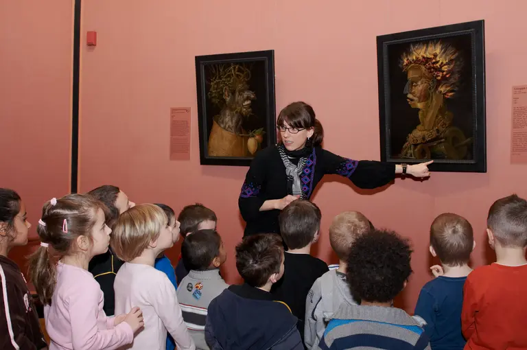 Eine Frau zeigt einer Gruppe von Kindern zwei Kunstwerke an einer Wand. Die Kinder hören aufmerksam zu, während sie auf die Bilder schauen. Die Wand ist rosa, und die Atmosphäre ist lernfreundlich und inspirierend.