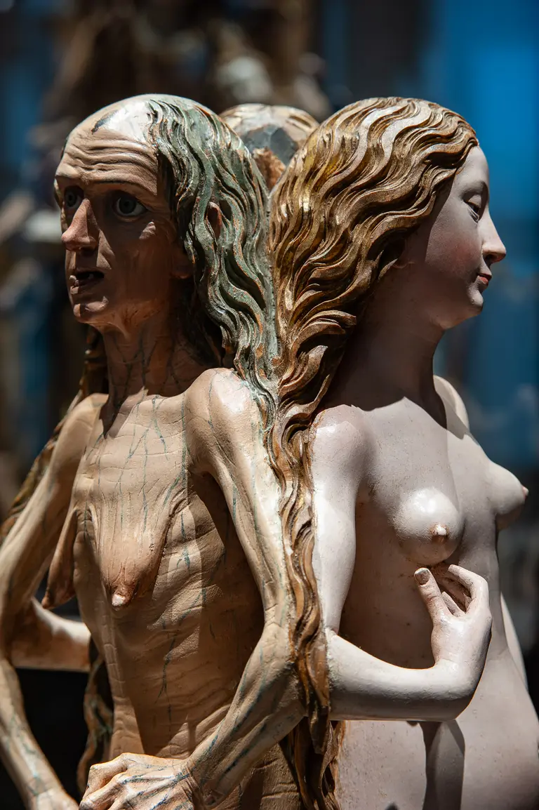 Die Skulptur zeigt eine männliche und eine weibliche Figur, die Rücken an Rücken stehen. Der Mann hat eine ausdrucksstarke Miene und lange, zerzauste Haare, während die Frau mit glatten Haaren und einem ruhigen Gesichtsausdruck dargestellt ist.