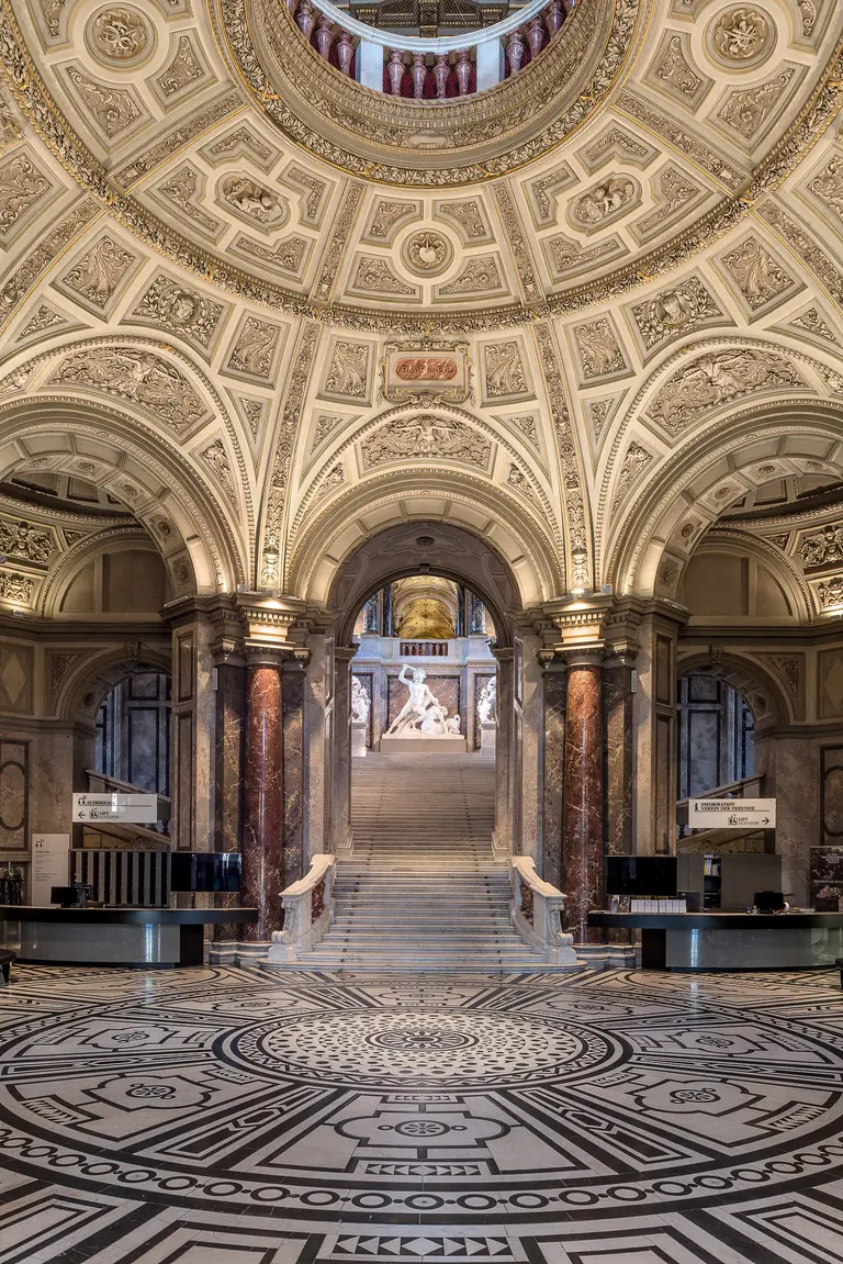 Impression des beeindruckenden Innenraums eines Museums mit einer kunstvoll gestalteten Decke und einem aufwendigen Bodenmuster. Im Hintergrund sind Stufen zu sehen, die zu einer Skulptur führen. Die Architektur strahlt Eleganz und historisches Flair aus.