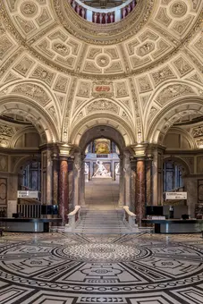 The image shows an elegant, grand interior of a museum hall. High ceilings with ornate decorations overlook a large staircase, leading to a statue at the top. The floor features a detailed geometric pattern, adding to the architectural beauty of the space.