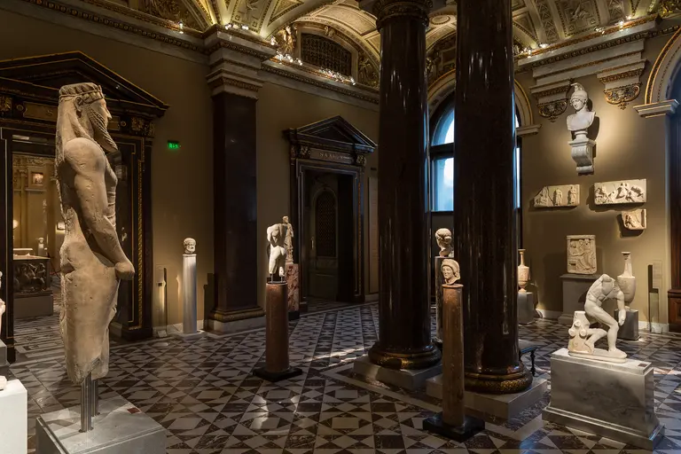 Das Bild zeigt einen Ausstellungsraum mit mehreren antiken Skulpturen und Reliefs. Auf marble Fußboden stehen Statuen, darunter eine große Figur und kleinere Skulpturen. Die Wände sind dekoriert, und große Säulen strukturieren den Raum, der ansprechend beleuchtet ist.